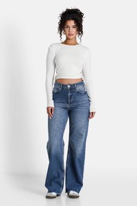 Top bianco a maniche lunghe con righe e lacci laterali, abbinato a jeans blu a vita alta e gamba larga. Il modello indossa scarpe da ginnastica grigie.