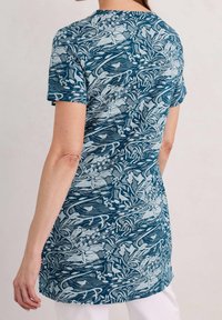 Túnica de manga corta en tejido azul con un patrón floral y abstracto detallado, con un corte recto y un dobladillo trasero ligeramente más largo.