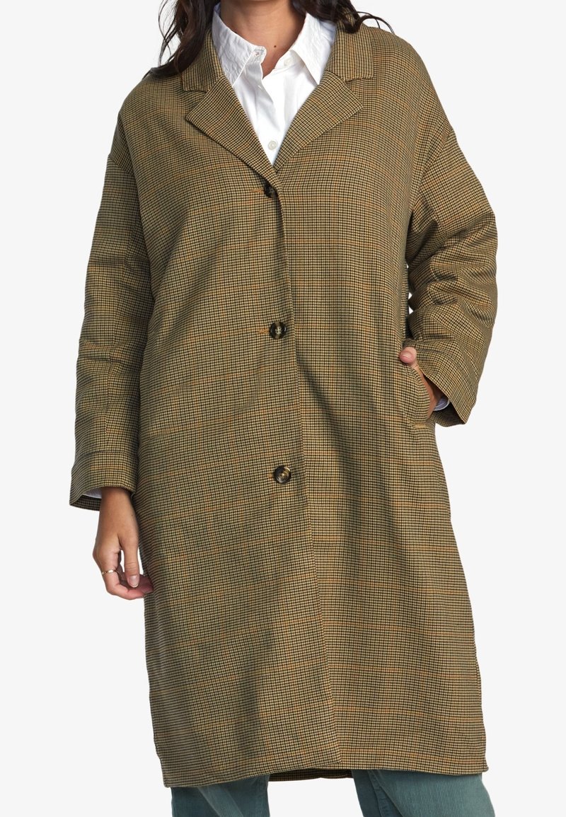 Femme portant un long manteau marron à carreaux avec des boutons, une chemise blanche à col, et un pantalon bleu sarcelle, debout avec une main dans la poche.