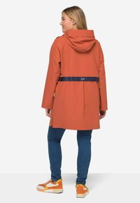 Oranssi vedenpitävä takki huppu ja tummansininen vyö, yhdistettynä sinisiin farkkuihin ja monivärisiin lenkkitossuihin. Silkkinen pinta ja keskipitkä muotoilu.