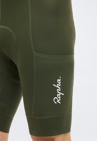 Ποδηλατικά σορτς σε ελιά πράσινο χρώμα με πλαϊνή τσέπη και λευκό λογότυπο Rapha στον μηρό, κοντινή λήψη στην κάτω κοιλιακή περιοχή και τα πάνω μέρη των ποδιών.