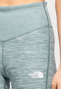 Leggings a vita alta in un tessuto verde opaco e testurizzato con motivo melange, dotati di un logo bianco sul lato sinistro.