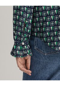 Blusa a maniche lunghe di colore blu navy con un motivo geometrico verde e bianco. Presenta un polsino arricciato con dettaglio a bottone e jeans in denim.