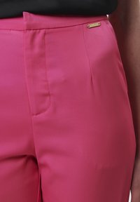 Aansluitende roze broek van gladde, lichte stof. Voorzien van een zijzak en een klein gouden logoaccent nabij de taille.