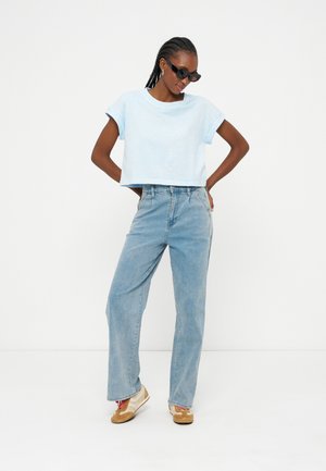Femme avec des cheveux tressés et des lunettes de soleil portant un t-shirt court bleu clair, un jean taille haute bleu clair et des baskets beige, posant les mains sur les hanches.