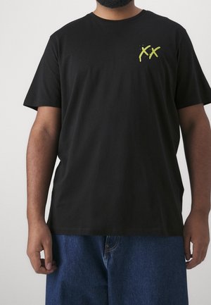 Mand iført sort T-shirt med gult dobbelt "X"-design på venstre bryst og blå jeans, stående mod en ensfarvet baggrund.