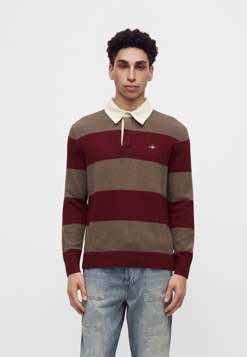 Jeune homme aux cheveux bouclés portant un pull rayé marron et bordeaux avec un col blanc et un jean bleu clair délavé.