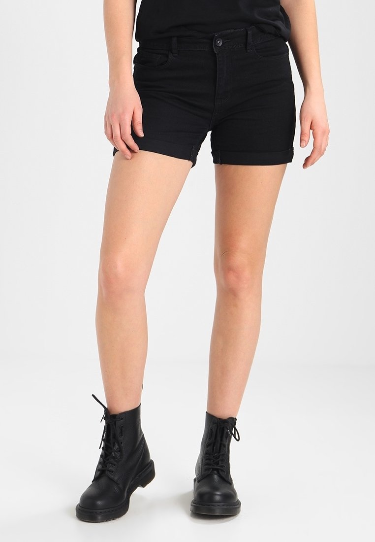 Shorts de denim negro con dobladillos enrollados, que presentan un diseño clásico de cinco bolsillos y un cierre frontal con botón. Se combinan con botas negras con cordones.