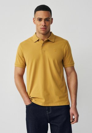 Młody mężczyzna w musztardowożółtej koszulce polo i ciemnoniebieskich dżinsach, stojący na tle jednolitego jasnoszarego tła.