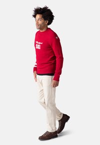 Roter Strickpullover mit "Ski"-Aufdruck, weißen Streifen an den Ärmeln, kombiniert mit cremefarbenen Hosen und braunen Wanderschuhen. Strukturieller, lässiger Outdoor-Stil.