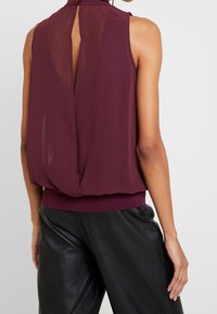 Blouse sans manches bordeaux avec un panneau arrière transparent, col haut et bas élastique. Le tissu semble léger et texturé, tombant doucement.