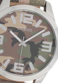Montre à motif camouflage avec boîtier en acier inoxydable, grands index horaires argentés et bracelet en tissu texturé de teintes neutres.
