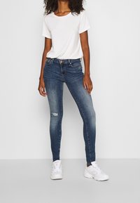 Mujer que lleva una camiseta blanca básica, jeans ajustados azules desgastados con un pequeño rasgón en una rodilla y zapatillas blancas, de pie contra un fondo gris.