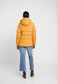 Gul puffjacka med huva, med horisontella quiltade sektioner, i kombination med ljus denim flare jeans och bruna klackstövlar.