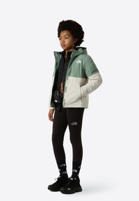 Veste à bloc de couleur verte et crème, superposition noire, leggings, chaussettes de marque, baskets noires, design épuré et accents fonctionnels.