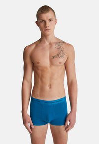 Uomo a torso nudo che indossa boxer blu con elastico "United Colors of Benetton", tatuaggio floreale sulla spalla e sul petto sinistro, sfondo neutro.