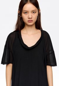 Musta lyhythihainen toppi, jossa on rosoinen kaula-aukko, kevyt kangas ja väljä malli, sekä hienovaraisia ommeltyylejä keskellä.