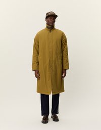 Dolga olivno zelena podložena bunda z visokim ovratnikom, dvema gumboma in sproščenim krojem, kombinirana z mornarsko modrimi hlačami in rjavimi čevlji.