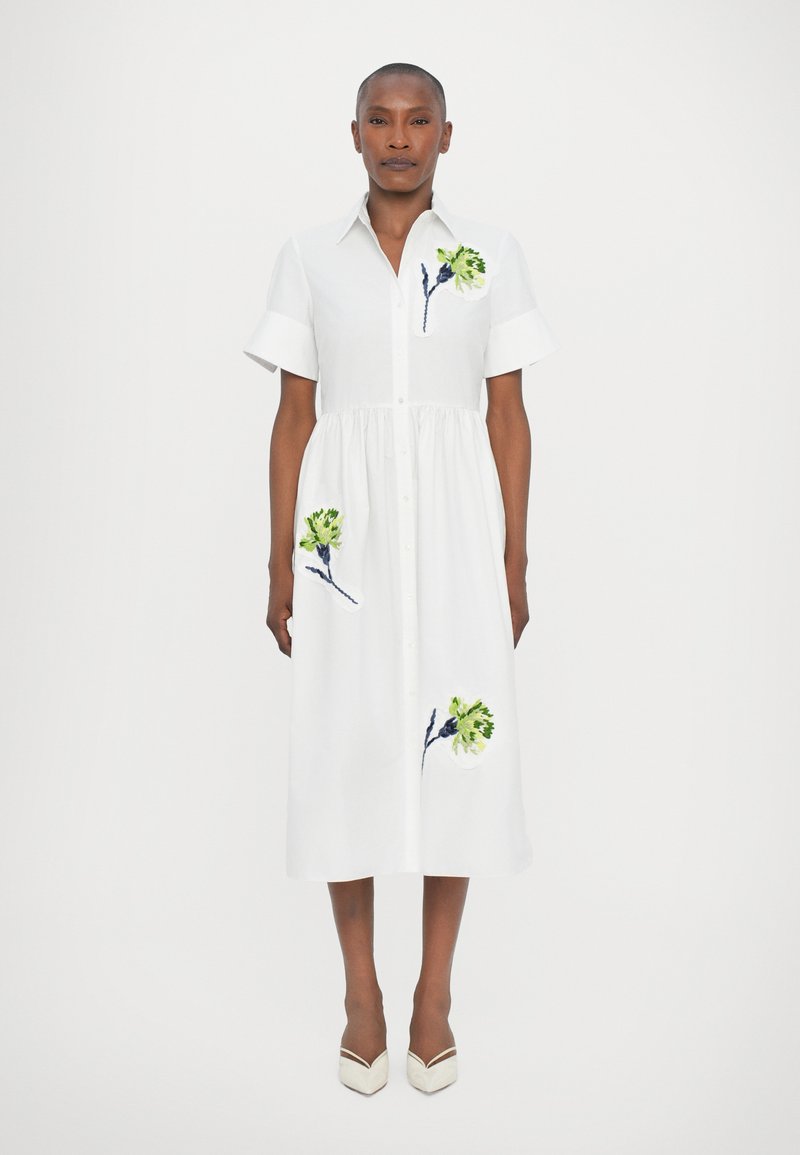 Abito bianco con bottoni e maniche corte, vita arricciata, con ricami floreali verdi e blu sulla parte frontale. Tessuto di cotone morbido.