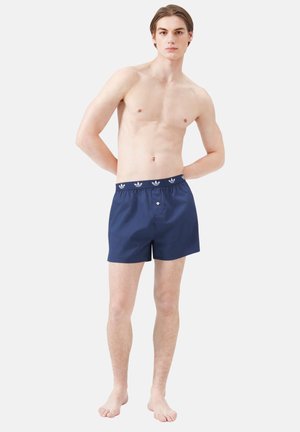 Jonge man staat op blote voeten en draagt marineblauwe boxershorts met een merknaam op de tailleband, tegen een effen witte achtergrond.