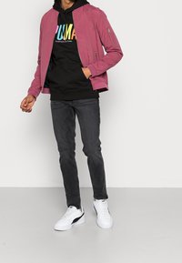 Giacca bomber rosa chiaro con zip frontale e tasche laterali, indossata sopra un felpa nera con un logo multicolore, abbinata a jeans scuri e sneakers bianche.