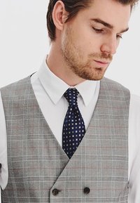 Szary kamizelka w kratę z czerwonymi akcentami, biała koszula i granatowy krawat w wzory. Posiada zapięcie na podwójne guziki oraz ostre kołnierzyki. Widoczna tekstura materiału.