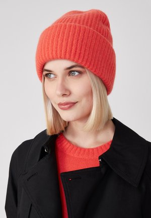 Strickmütze in leuchtendem Korallenrot, mit gerippter Textur und lockerer Passform. Getragen mit einem schwarzen Mantel und einem orangefarbenen Pullover darunter.