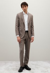 Abito di colore marrone chiaro con tessuto textures, giacca monopetto e pantaloni coordinati. Indossato con una camicia bianca e scarpe di pelle nere.