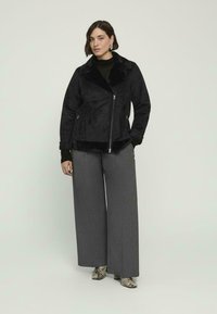 Giacca in camoscio nero con ampio colletto, chiusura con zip e finitura strutturata, abbinata a pantaloni grigi a gamba larga e tacchi con stampa a serpente.