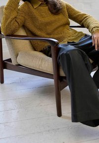 Hellbrauner, strukturierter Pullover mit sichtbarem Kragen, kombiniert mit weit geschnittenen schwarzen Hosen, sitzend auf einem Holzstuhl mit beigem Polster.