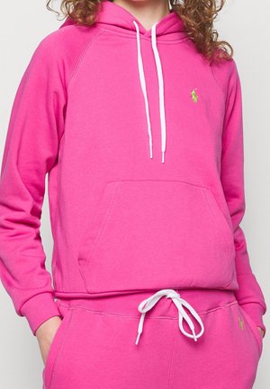 Persoon draagt felroze hoodie en bijpassende trainingsbroek met witte aantrekkoorden en klein groen geborduurd logo op de borst en broek.