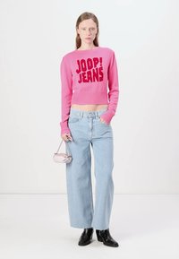 Pink cropped sweater with raised "JOOP! JEANS" text, paired with light blue wide-leg jeans. Model holds a small pink handbag and wears black boots.