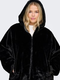 Young woman with blonde hair wearing a black plush zip-up hooded jacket with hands in pockets against a plain background.