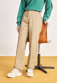 Pantaloni beige con un design a gamba dritta, caratterizzati da una texture liscia e dettagli nella cucitura laterale, abbinati a sneakers di colore chiaro.