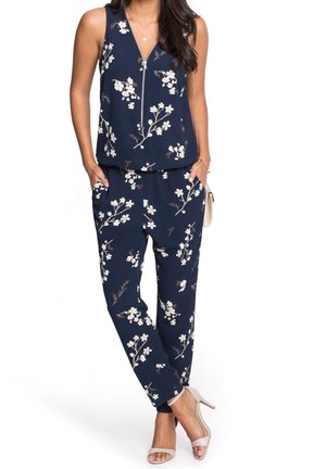 Combinaison florale bleu marine avec un décolleté en V, présentant des motifs de fleurs blanches, une taille élastique et des poches latérales. Associée à des talons nude.