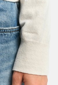 Heller beigefarbener, gerippter Pulloverbund vor blauer Jeans, der eine weiche Textur mit einem auffälligen Strickmuster und minimalen Nähten präsentiert.
