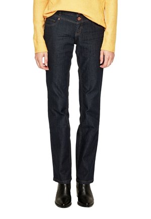 Femme portant un jean droit bleu foncé, un pull jaune et des bottines noires cloutées argent, debout avec les mains détendues le long du corps.