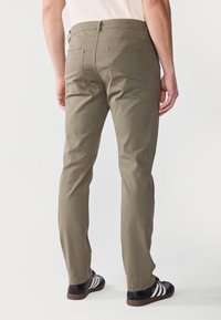 Khaki hlače s naravnim krojem in dvema zadnjima žepoma. Material ima gladko teksturo. Oblečene z črnimi čevlji in belimi detajli.