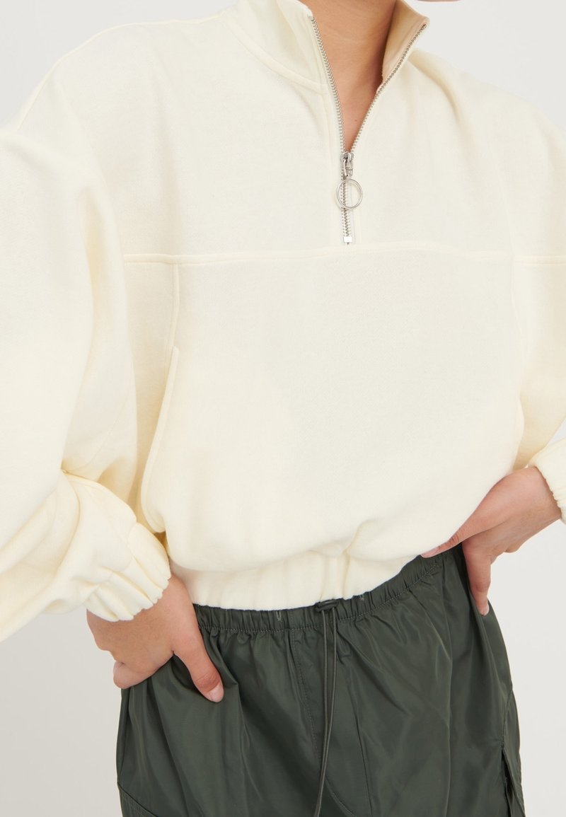 Personne portant un pull à fermeture éclair courte crème avec des poignets élastiques et un pantalon à taille élastique vert foncé, les mains posées sur les hanches.