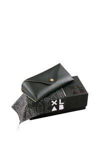 Custodia per occhiali da sole in pelle nera con chiusura a scatto, posizionata su una scatola nera con un motivo bianco di lettere e forme. Panno per la pulizia visibile.