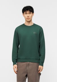 Νεαρός άνδρας με κοντά μαύρα μαλλιά, φορώντας σκούρο πράσινο φούτερ και καφέ παντελόνι cargo, σε απλό λευκό φόντο.