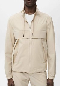 Beige letvægts zip-up jakke med høj krave, frontlommer og snoretræk. Har en slank, glat tekstur og minimalistisk design.