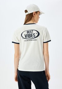 Cremefarvet t-shirt med marineblå kant har et stort grafisk tryk med teksten "BEST VIBES" på ryggen, med yderligere tekst nedenunder.