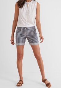 Top blanco sin mangas con detalles de ojal y cuello con cordón, combinado con pantalones cortos a rayas azul y blanco, y sandalias planas marrones.