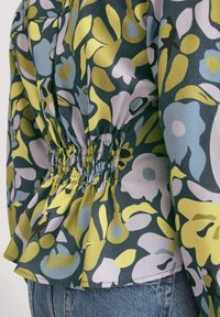 Primo piano di una blusa floreale colorata con vita arricciata indossata con jeans blu, che mette in evidenza il motivo del tessuto e il dettaglio arricciato.
