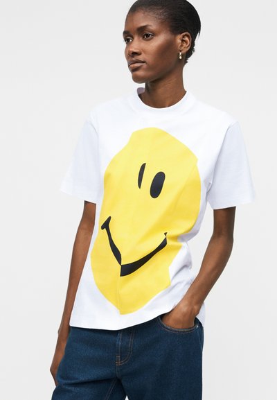 Γυναίκα με λευκό t-shirt με μεγάλο κίτρινο χαμογελαστό γραφικό, συνδυασμένο με μπλε τζιν, στέκεται με το ένα χέρι στην τσέπη.