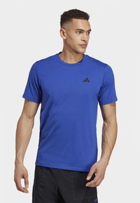 Muž v modrom tričku Adidas s krátkym rukávom a čiernych šortkách, pozerajúci sa do strany s neutrálnym výrazom tváre na jednoduchom pozadí.