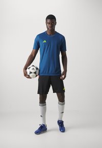 Blue athletic jersey with green accents, black shorts, blue soccer cleats, and white socks with black stripes. Player holding a soccer ball.