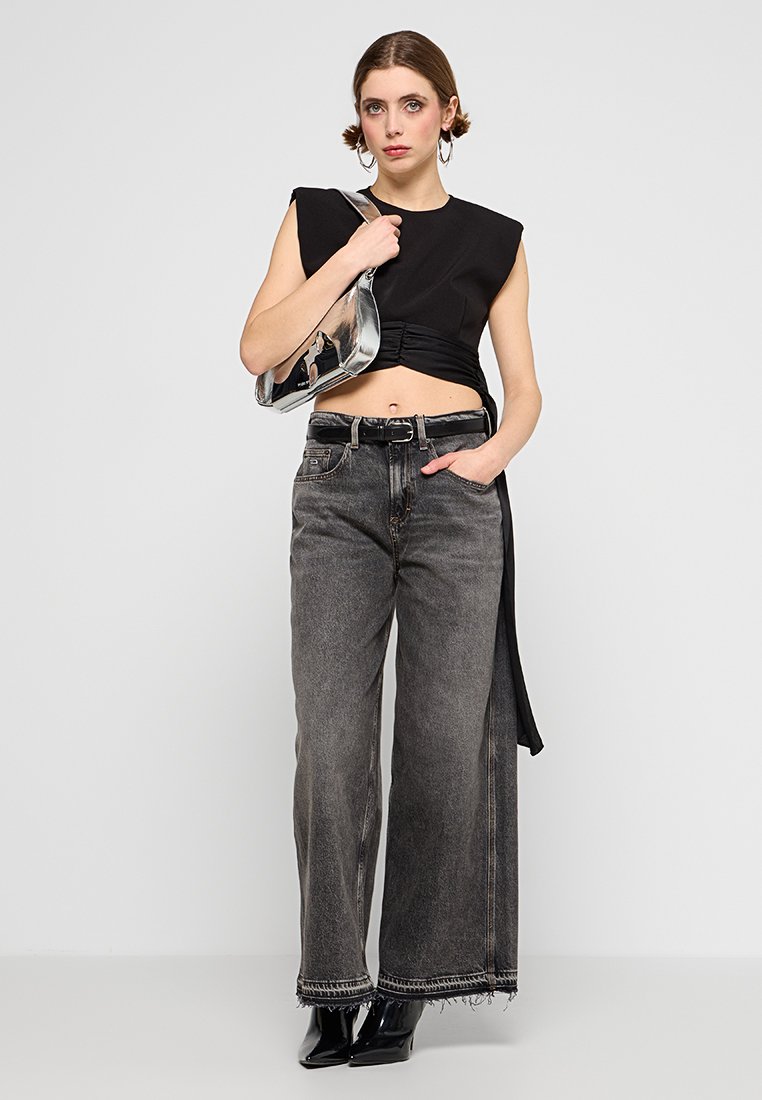 Schwarzes Crop-Top mit strukturierten Schultern, kombiniert mit hochtaillierten, weit geschnittenen grauen Jeans mit fransigen Säumen und einem schwarzen Gürtel. Silberne Handtasche.