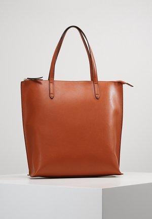 Braune Ledertasche mit zwei Schulterriemen und einem oberen Reißverschluss, steht aufrecht auf einem weißen Podest vor einem schlichten Hintergrund.